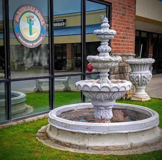 Three-tiered concrete fountain in front of a store window with a logo and grass.