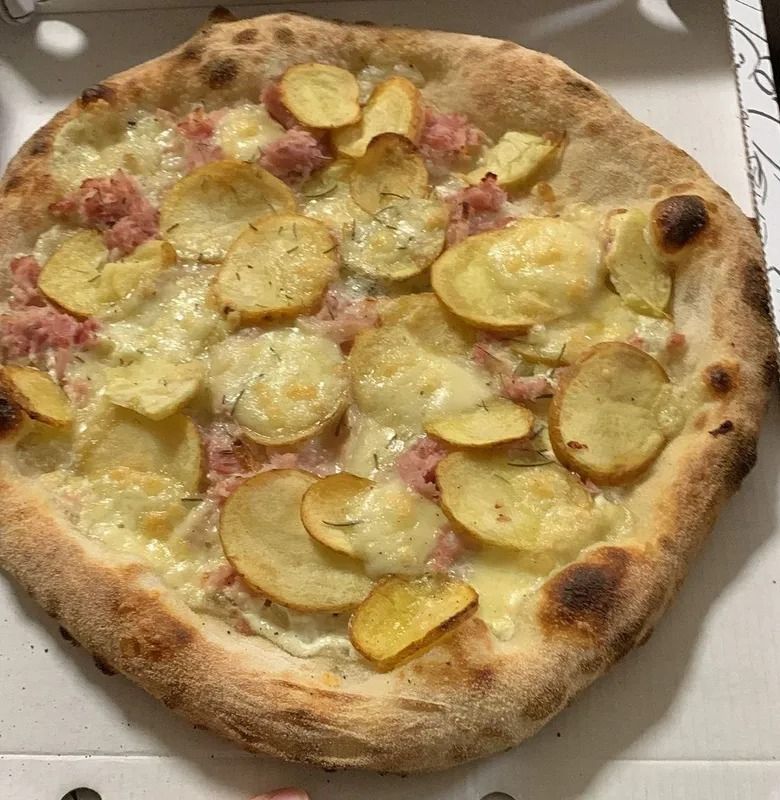 Una pizza rotonda con patate sopra, servita in una scatola bianca, con formaggio fuso e una crosta dorata.
