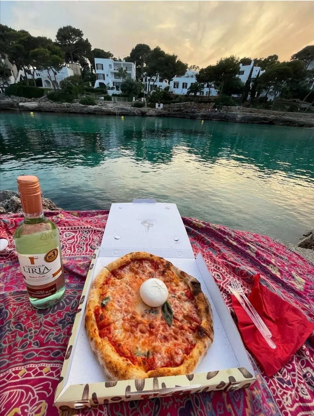 Pizza e una bottiglia su una coperta a fantasia in riva al mare turchese al tramonto, con le case che si stagliano all'orizzonte.