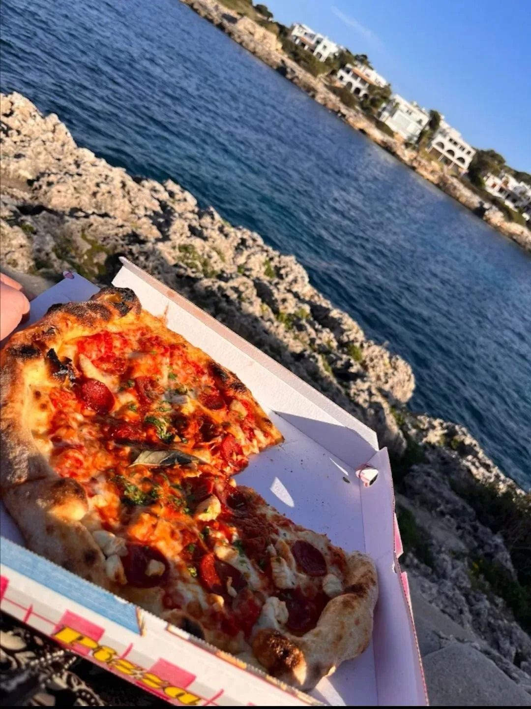Pizza in scatola tenuta in mano accanto a una costa rocciosa con acqua blu e edifici sullo sfondo.