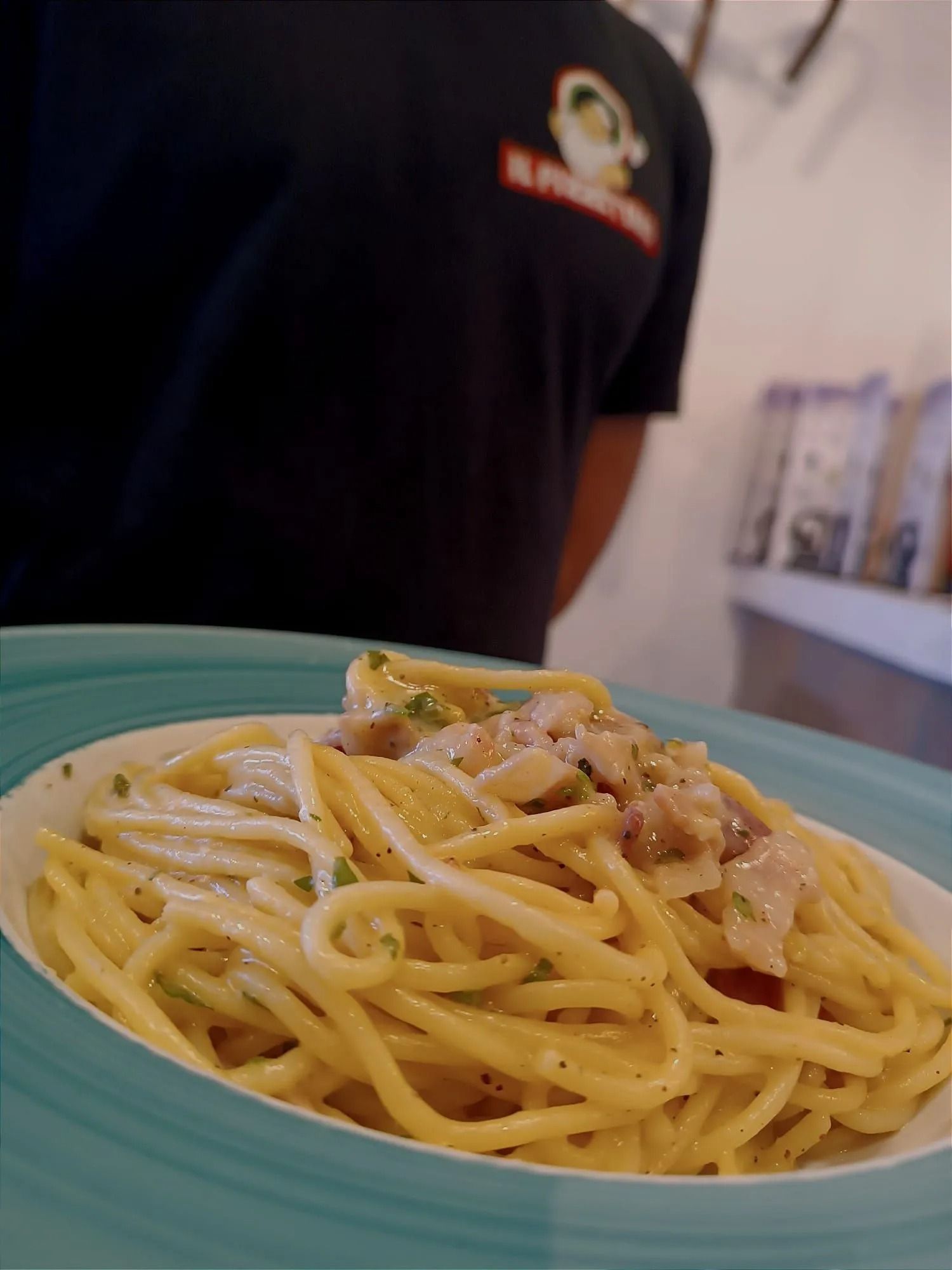 In primo piano, un piatto di spaghetti su un vassoio turchese, con una persona in piedi sullo sfondo.