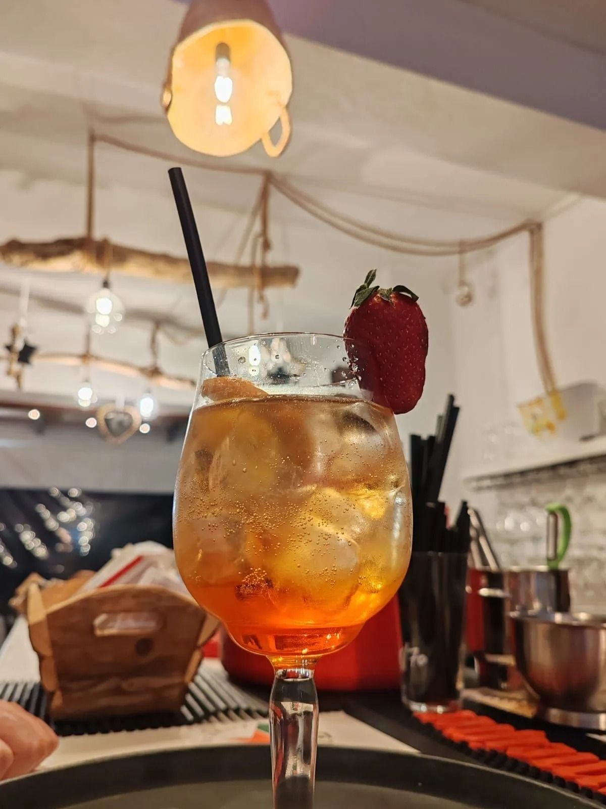 Cocktail all'arancia con guarnizione di fragola e cannuccia su un tavolo da bar, illuminato da calde luci sospese.