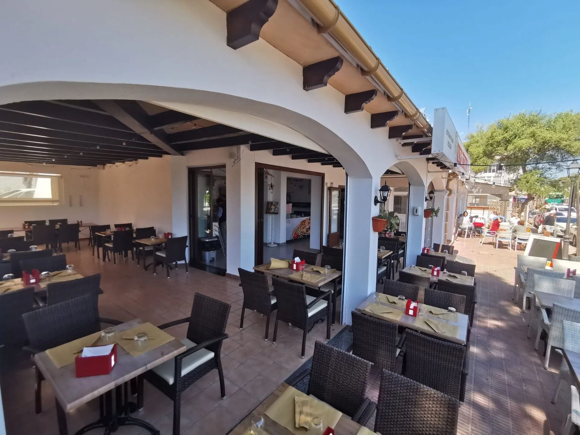 Terrazza esterna del ristorante con tavoli e sedie in vimini sotto archi bianchi, affacciata su una strada soleggiata.