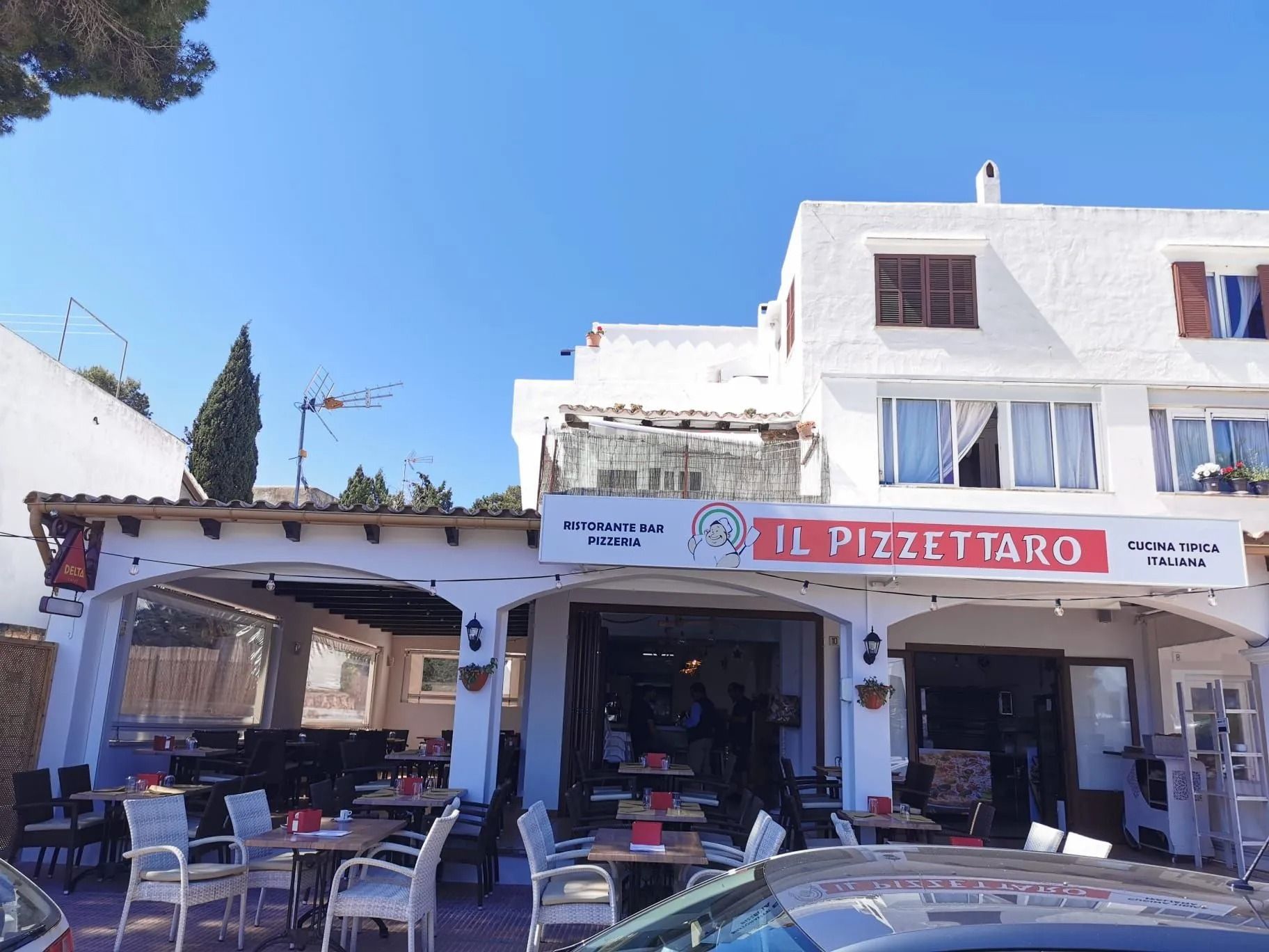 Caffè all'aperto con sedie blu e tenda da sole rossa di fronte a un edificio bianco sotto un cielo limpido.