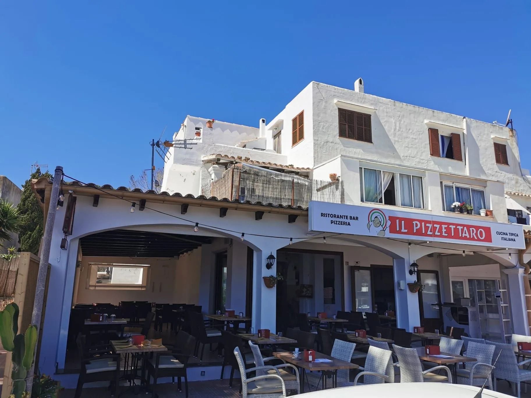Ristorante all'aperto con edifici bianchi, cielo azzurro e tavoli sotto un patio ombreggiato.