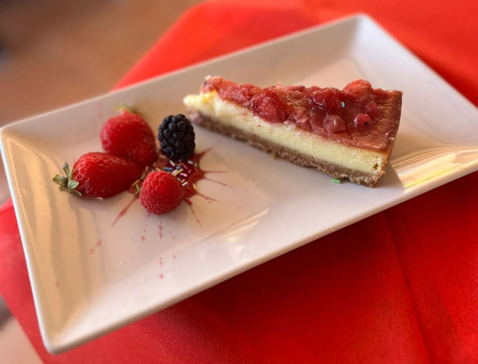 Una fetta di cheesecake alla fragola con lamponi e more su un piatto bianco sopra una tovaglia rossa.