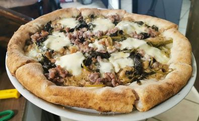 Primo piano di una pizza con formaggio e carne su un vassoio, con una crosta spessa e vari condimenti, in un ambiente rustico.