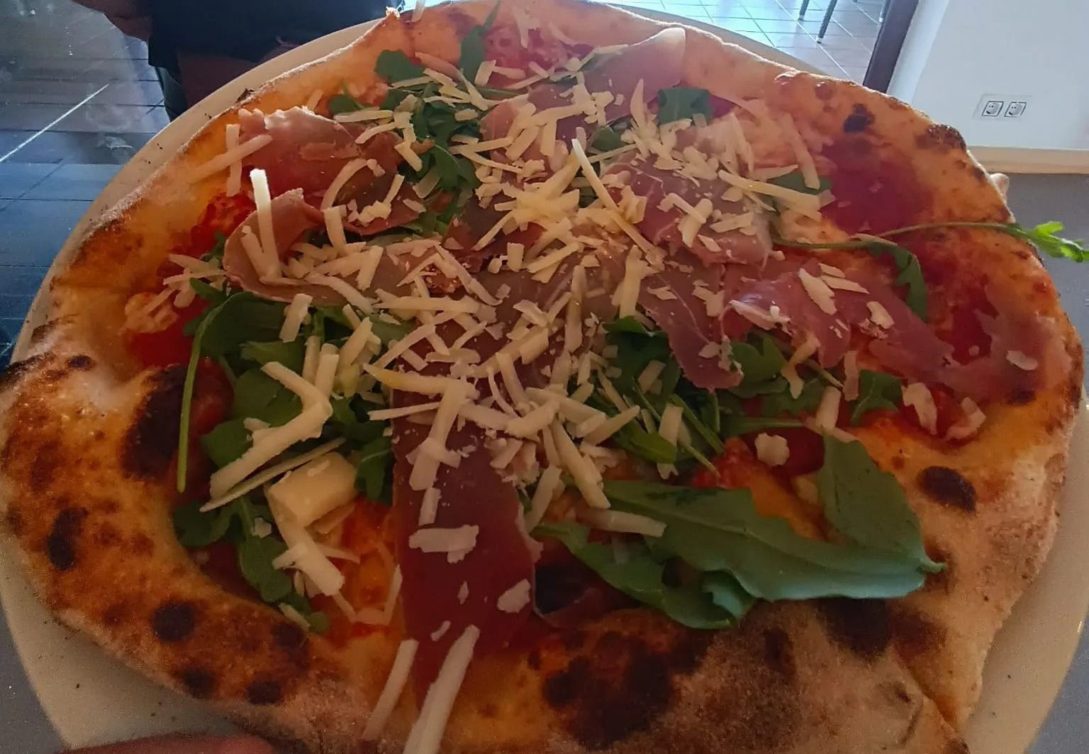 Una pizza condita con rucola, formaggio grattugiato e salumi, servita su un piatto bianco.