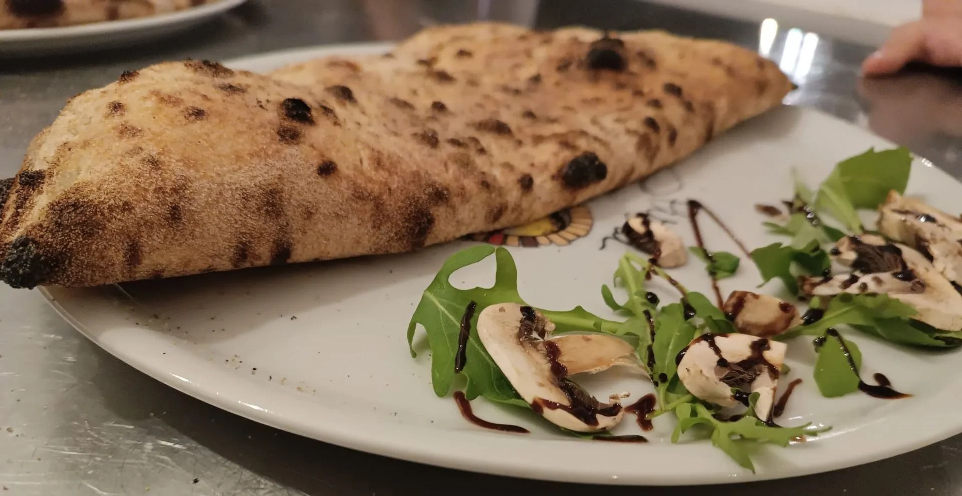 Calzone cotto nel forno a legna su un piatto, accompagnato da una piccola insalata di rucola con un filo di aceto balsamico.