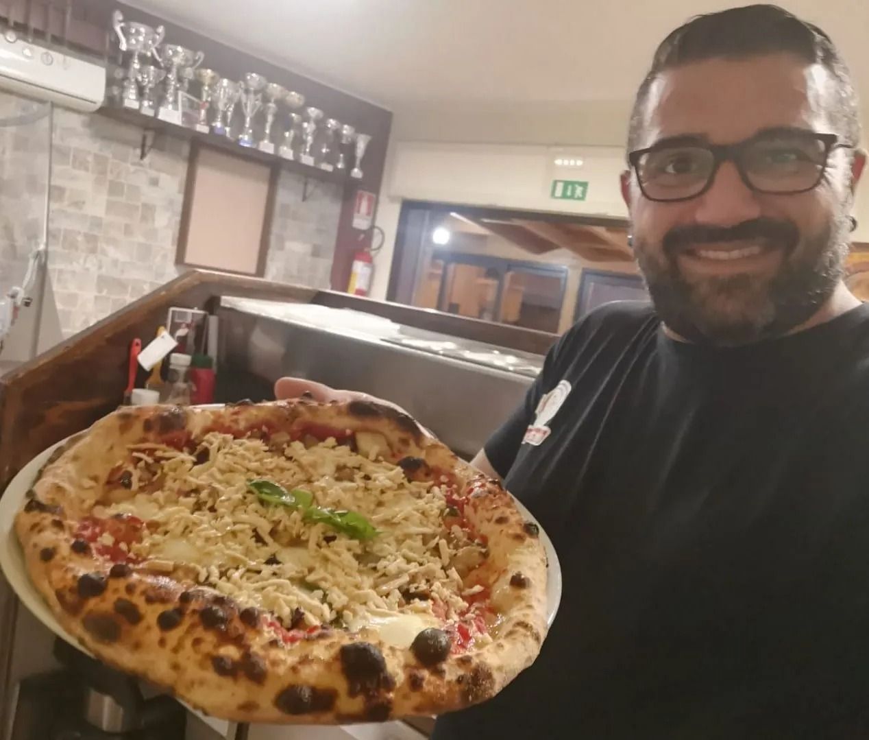 Un uomo sorridente tiene in mano una grande pizza in un ristorante, con un ambiente interno accogliente e luci a parete alle sue spalle.