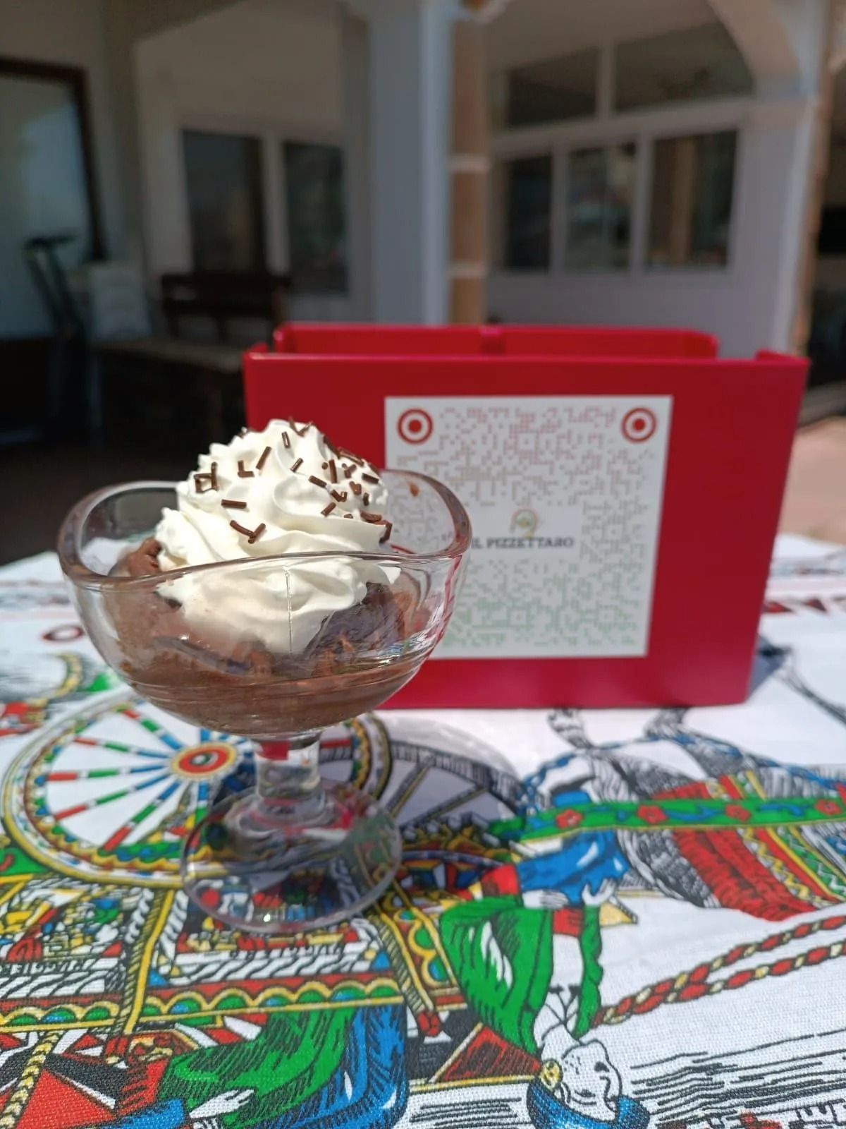 Coppa di gelato in una coppetta di vetro su una tovaglia colorata, con una ricetta scritta su un cartoncino rosso sullo sfondo.
