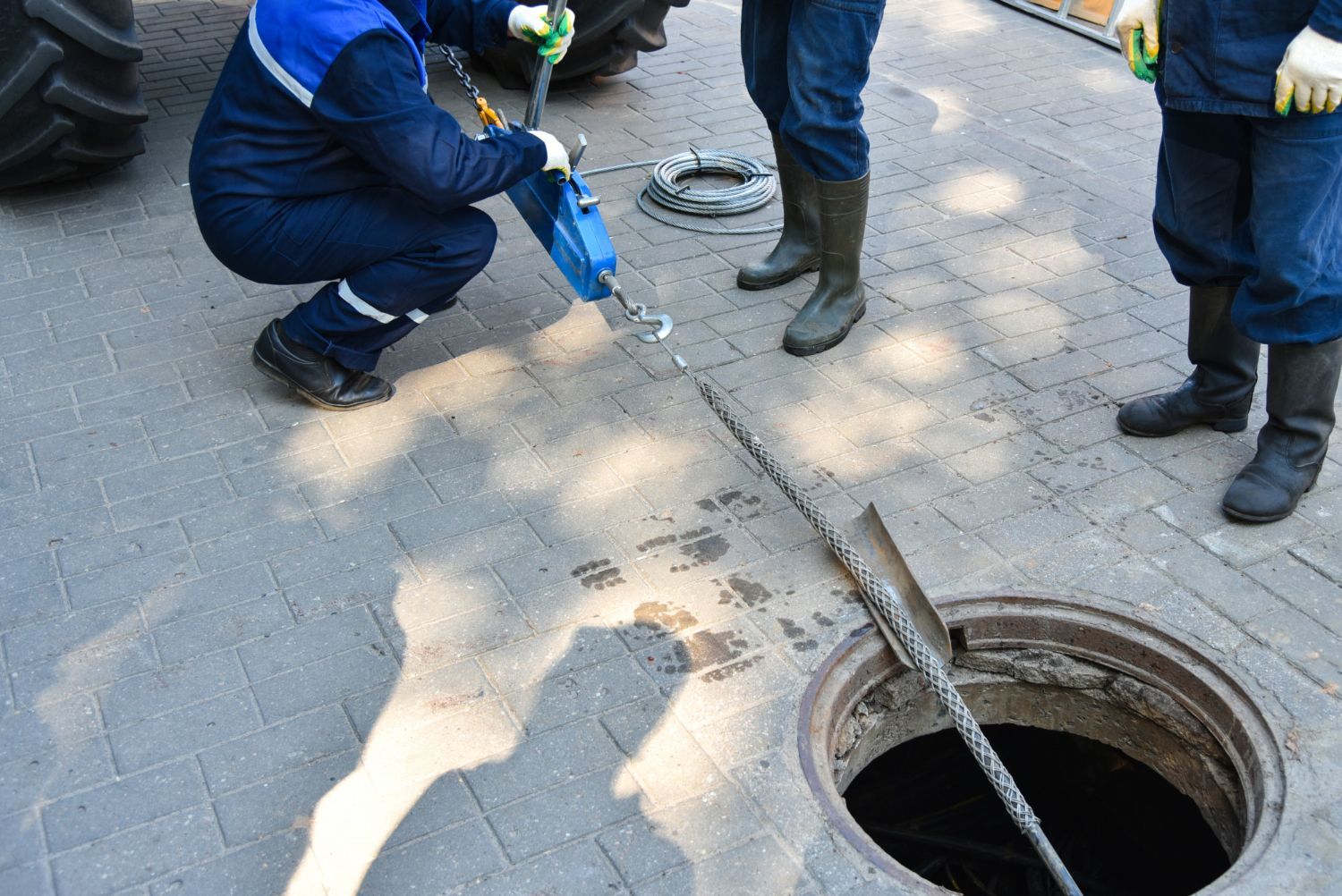 Workers inspecting a manhole; one uses a tool, others wear boots, outdoors.