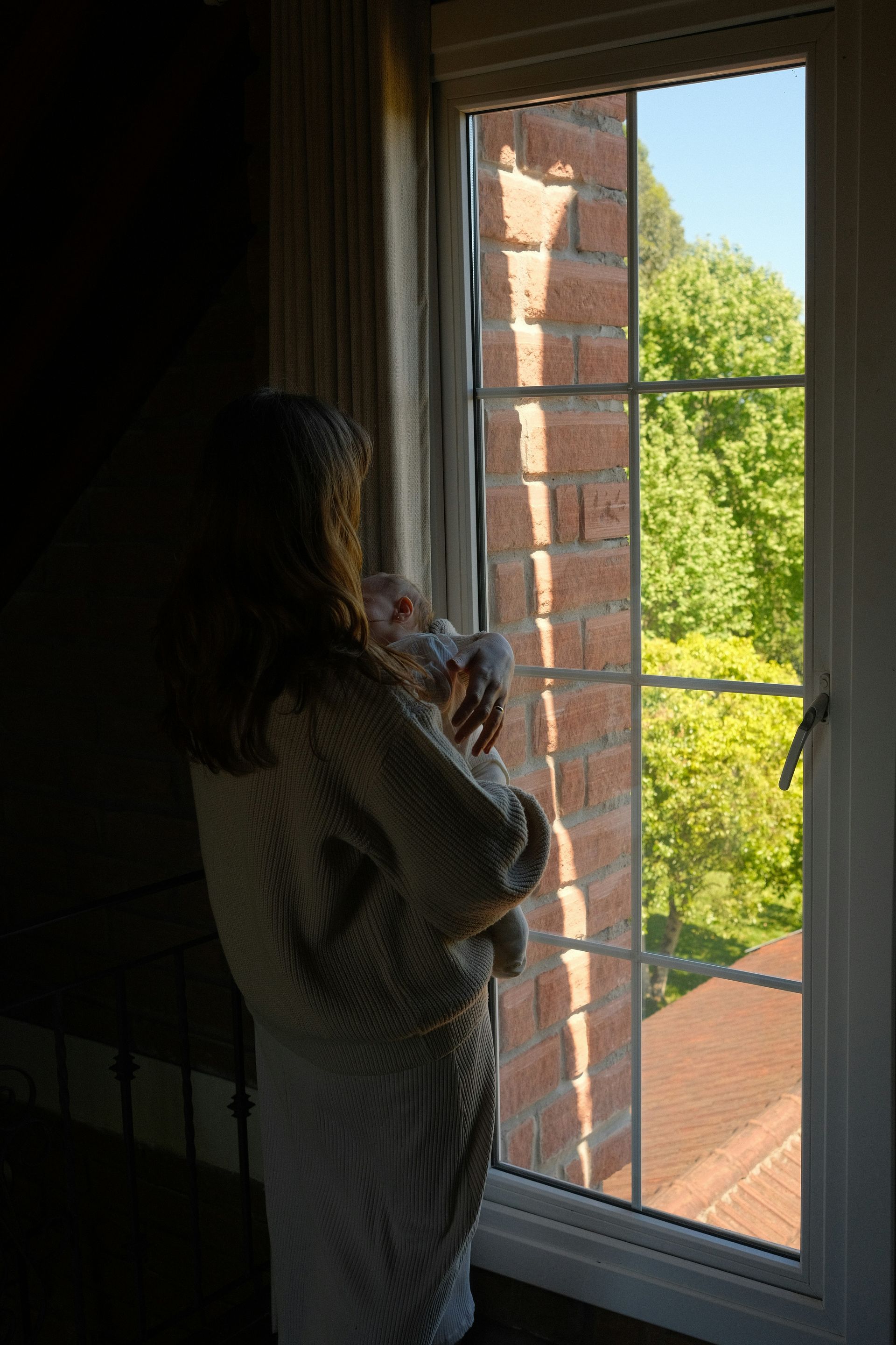 Woman holding a baby looks out a window at a brick wall and trees.