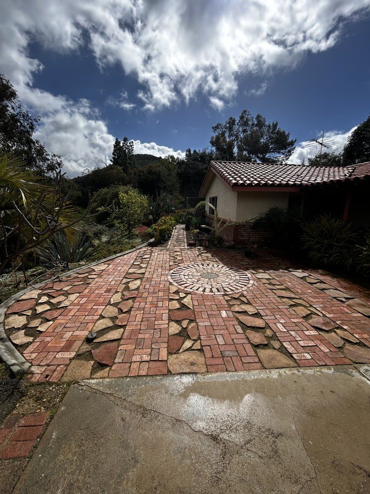 Brick driveway leading to a house under a cloudy sky, with trees and shrubs along the sides.