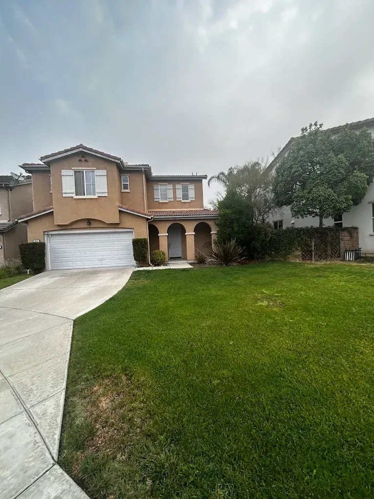 Two-story tan house with a garage and arched entry, fronted by a large green lawn under a cloudy sky.