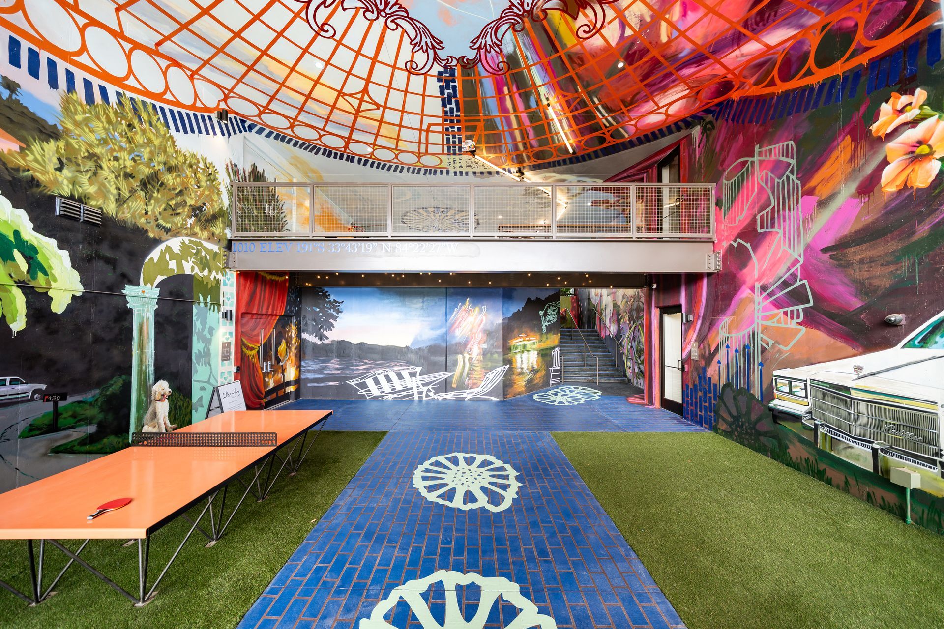 A colorful indoor recreational space with a ping pong table, artificial grass, a blue pathway, and vibrant mural walls.