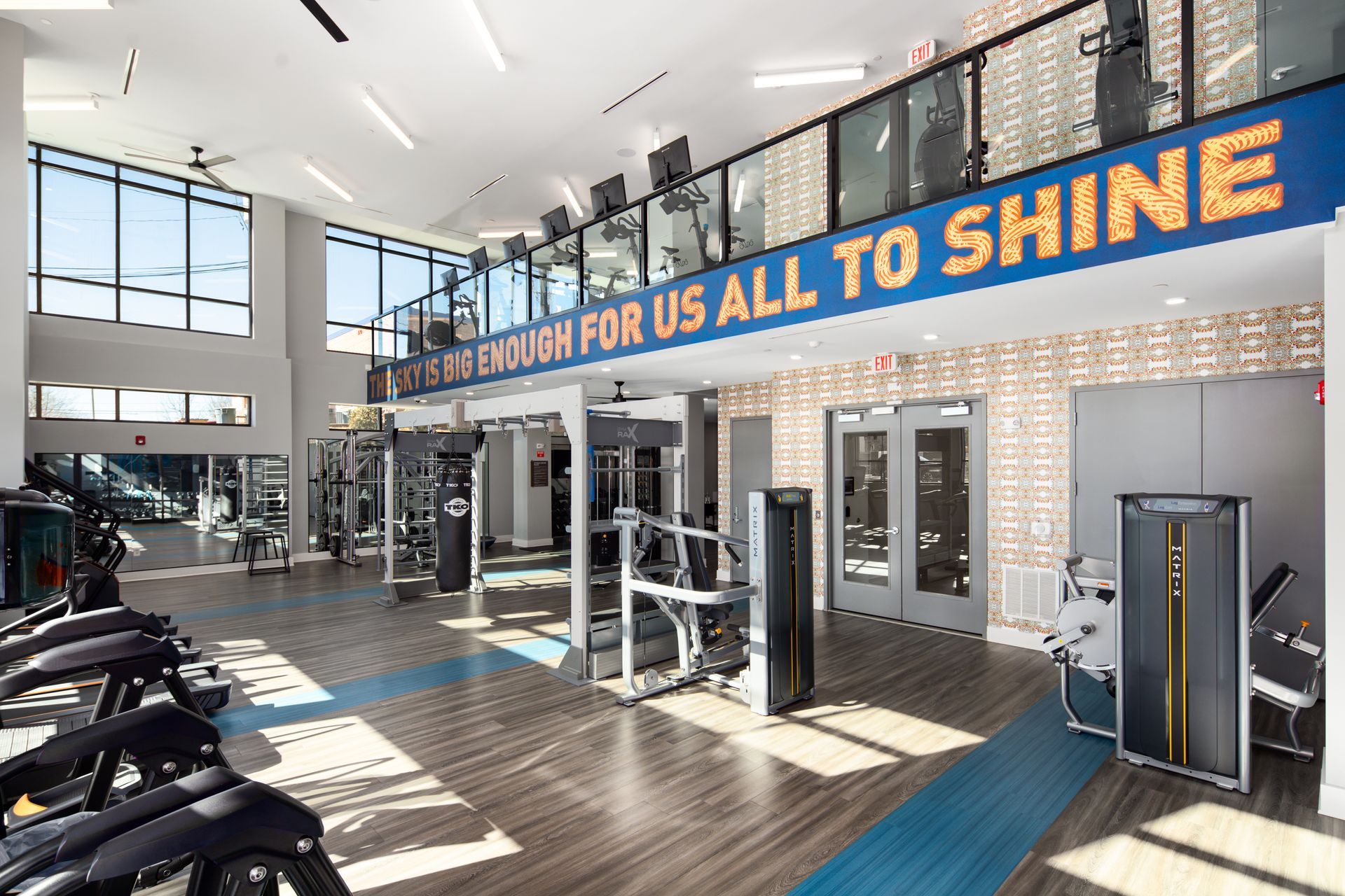 Modern gym interior with exercise machines, wood-look flooring, and a blue balcony sign reading 