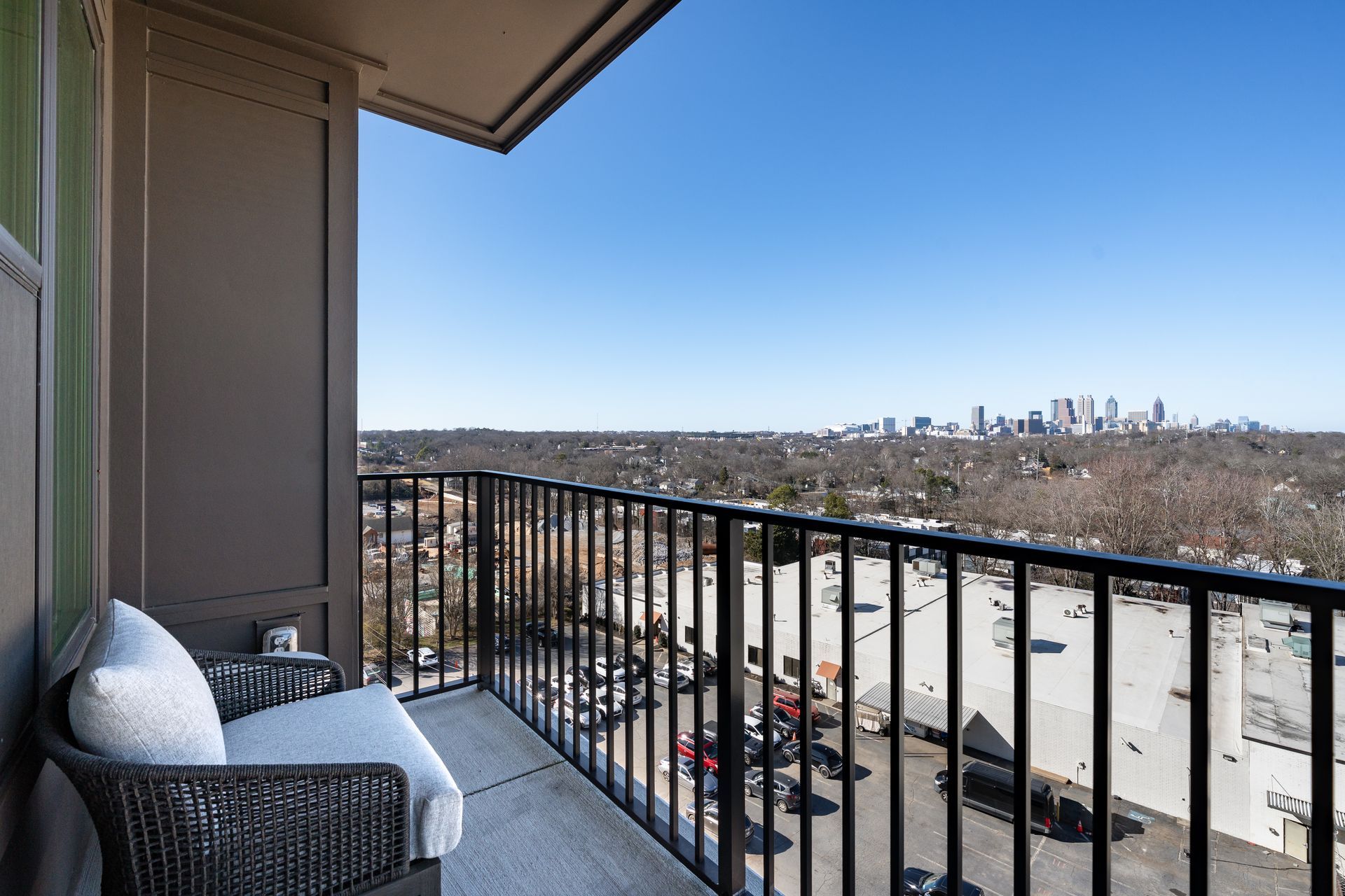 A balcony with a woven chair overlooking a suburban area and a distant city skyline under a clear blue sky.