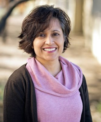Smiling person with shoulder-length dark hair wearing a pink cowl-neck top and dark cardigan outdoors.