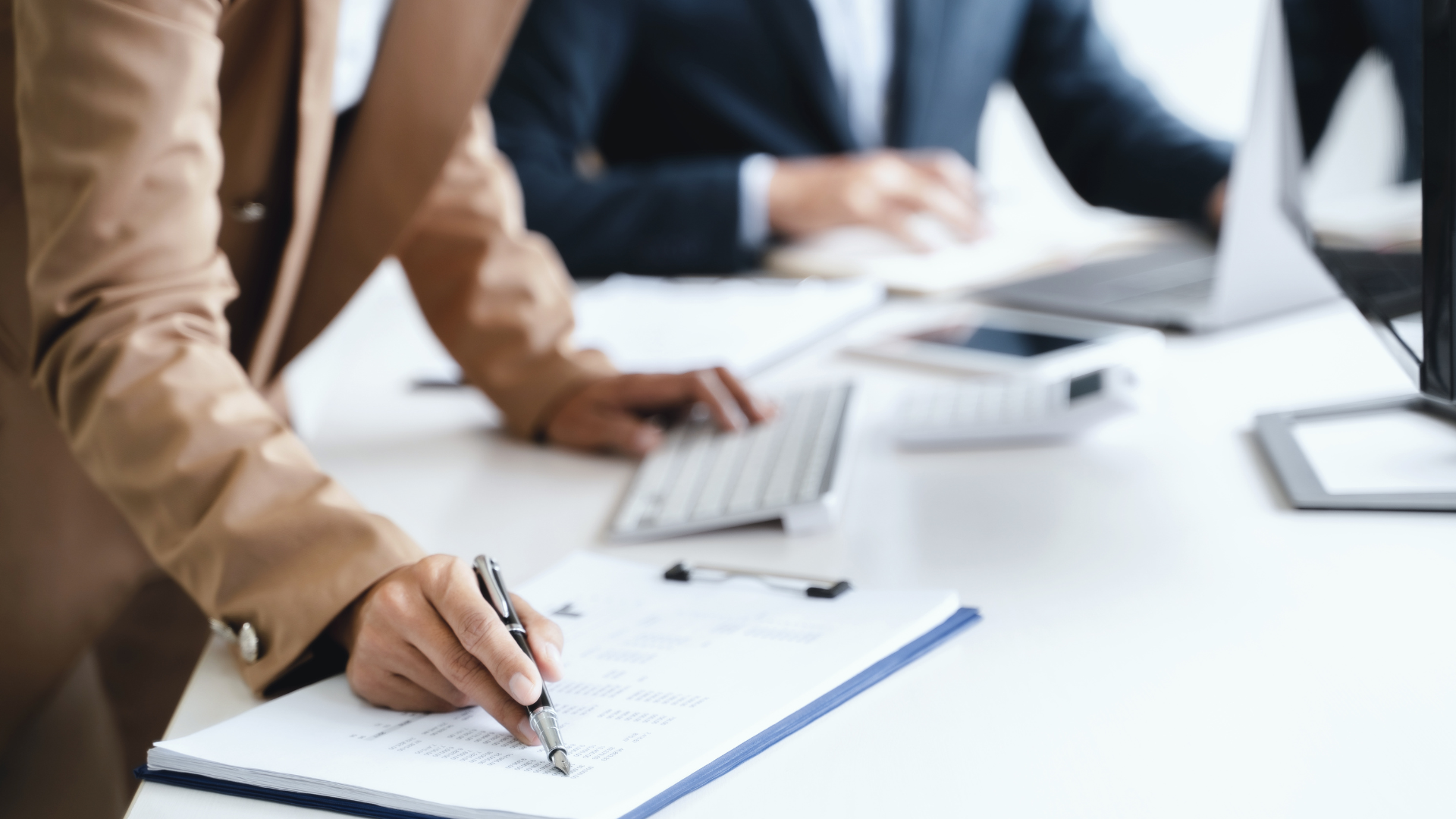 Hands writing notes at a conference table with laptops and papers in a bright office meeting room