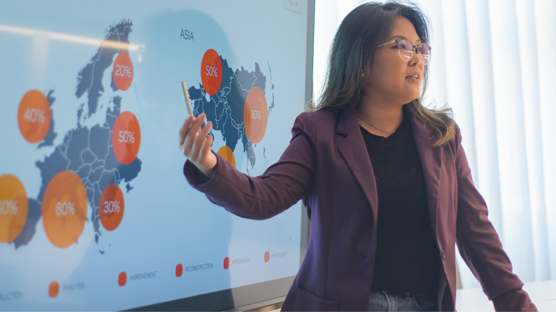 Presenter in a maroon blazer gesturing at a world map dashboard on a large screen.