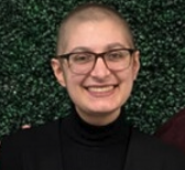 Smiling person with glasses and black turtleneck in front of a leafy green backdrop