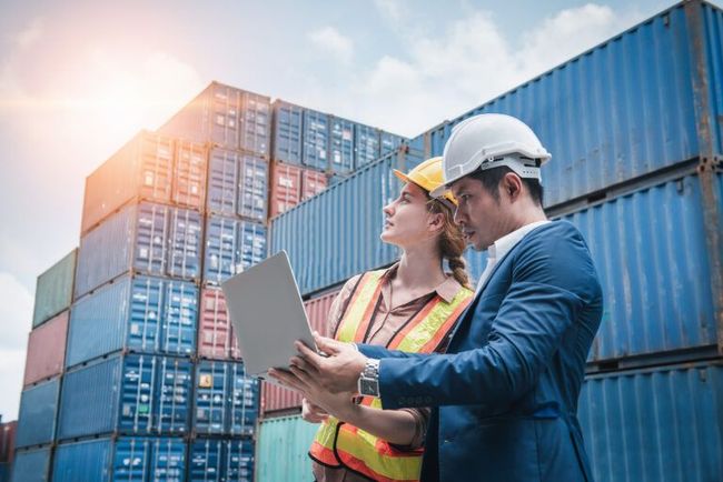 Two workers in hard hats review plans beside stacked shipping containers in a port.