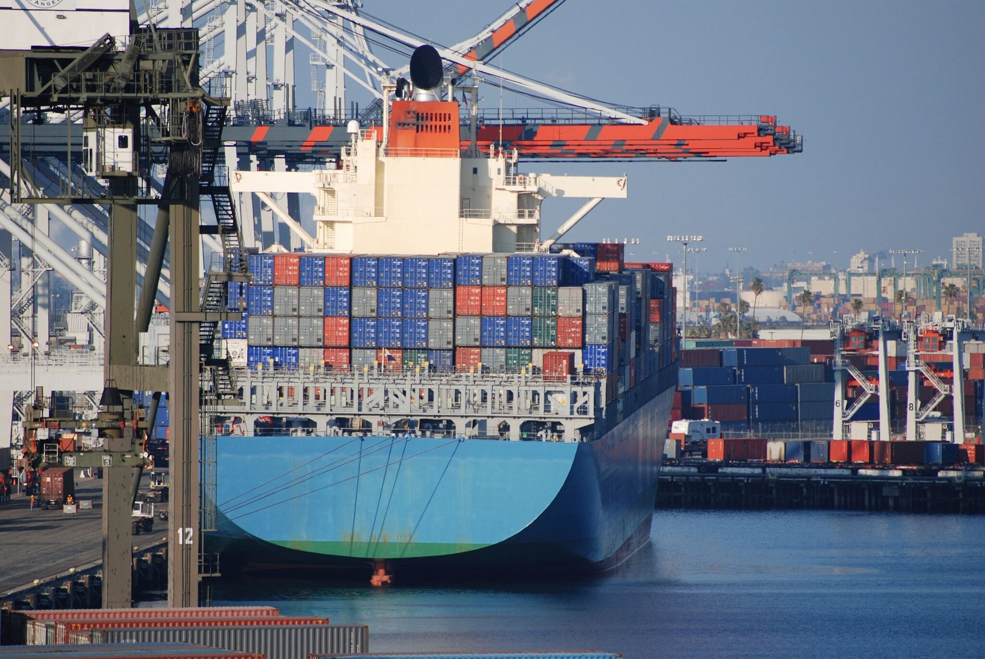 Cargo ship loaded with containers in a busy port, seen from the stern under cranes.