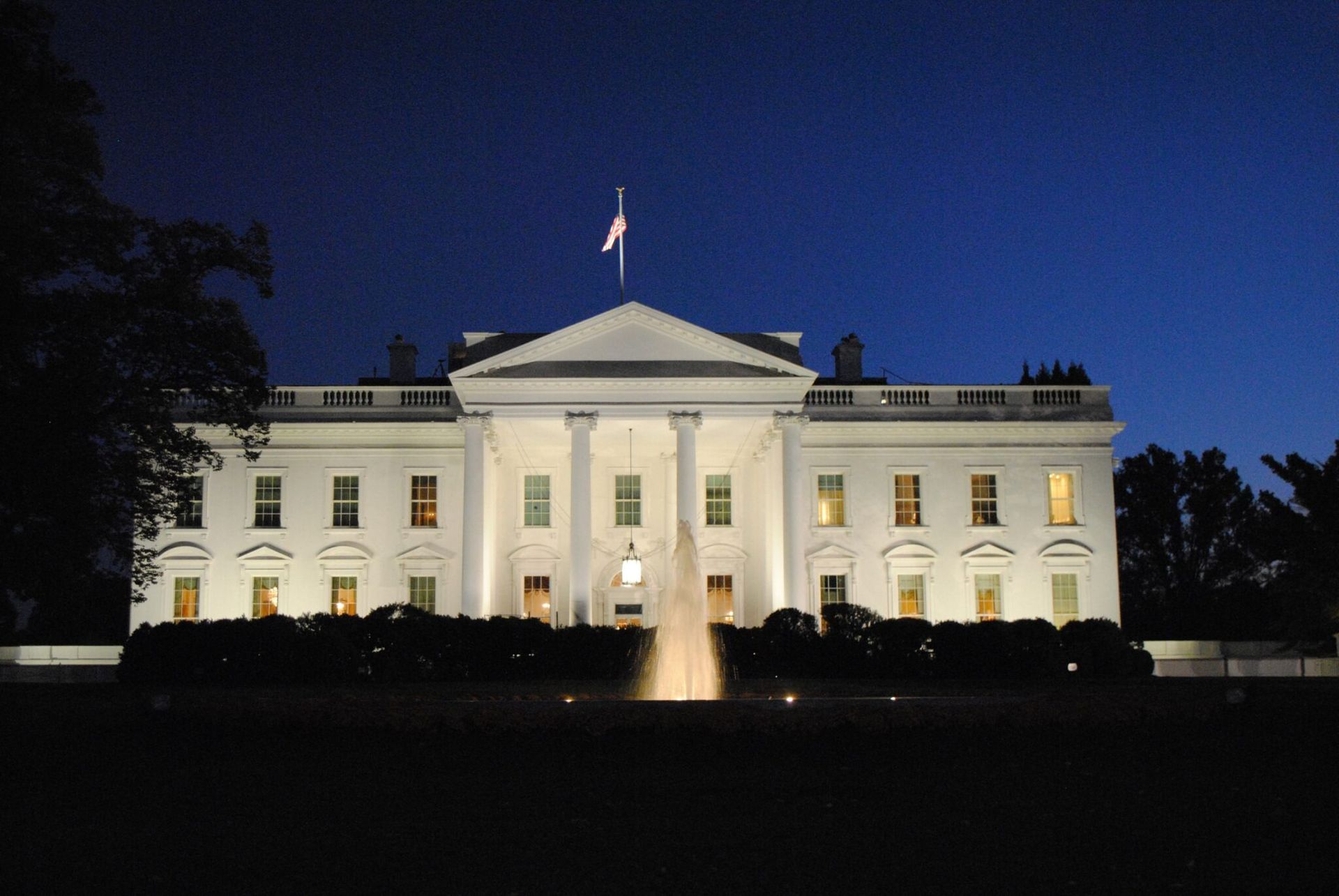 The White House at night, illuminated against a dark sky with the fountain in front.