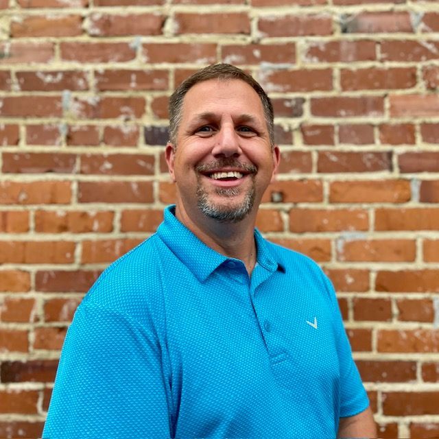 Smiling man in a blue polo shirt standing against a brick wall.