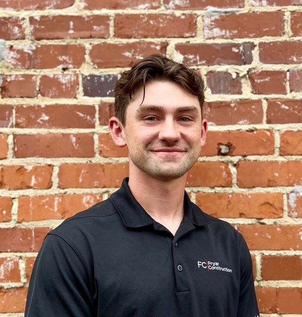 Man with a slight smile wearing a black polo shirt, sitting in front of a green wall.