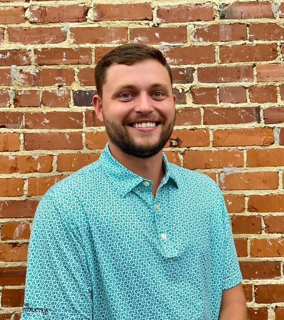 Man with beard in black polo shirt smiles in front of a green wall.