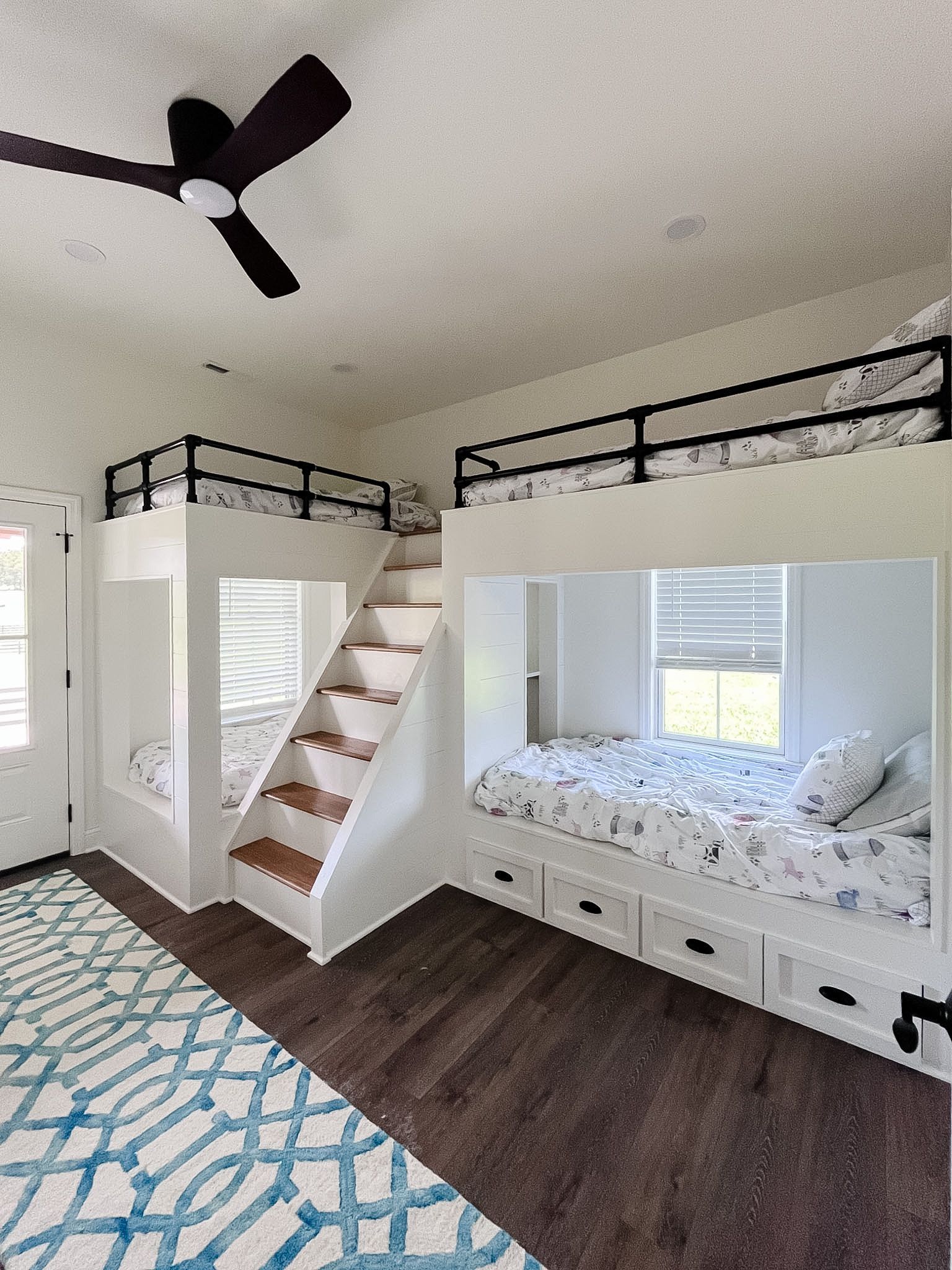 Bright loft-style bedroom with bunk beds, stairs, dark wood floor, blue patterned rug, and ceiling fan