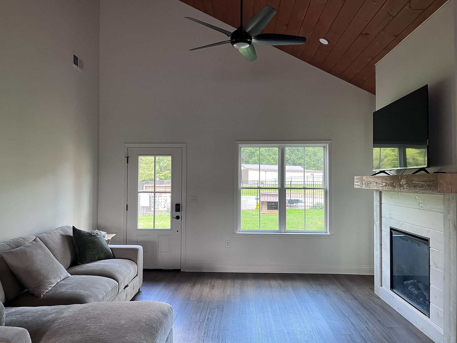 Bright living room with gray sofa, fireplace, large window, and vaulted wood ceiling with ceiling fan.