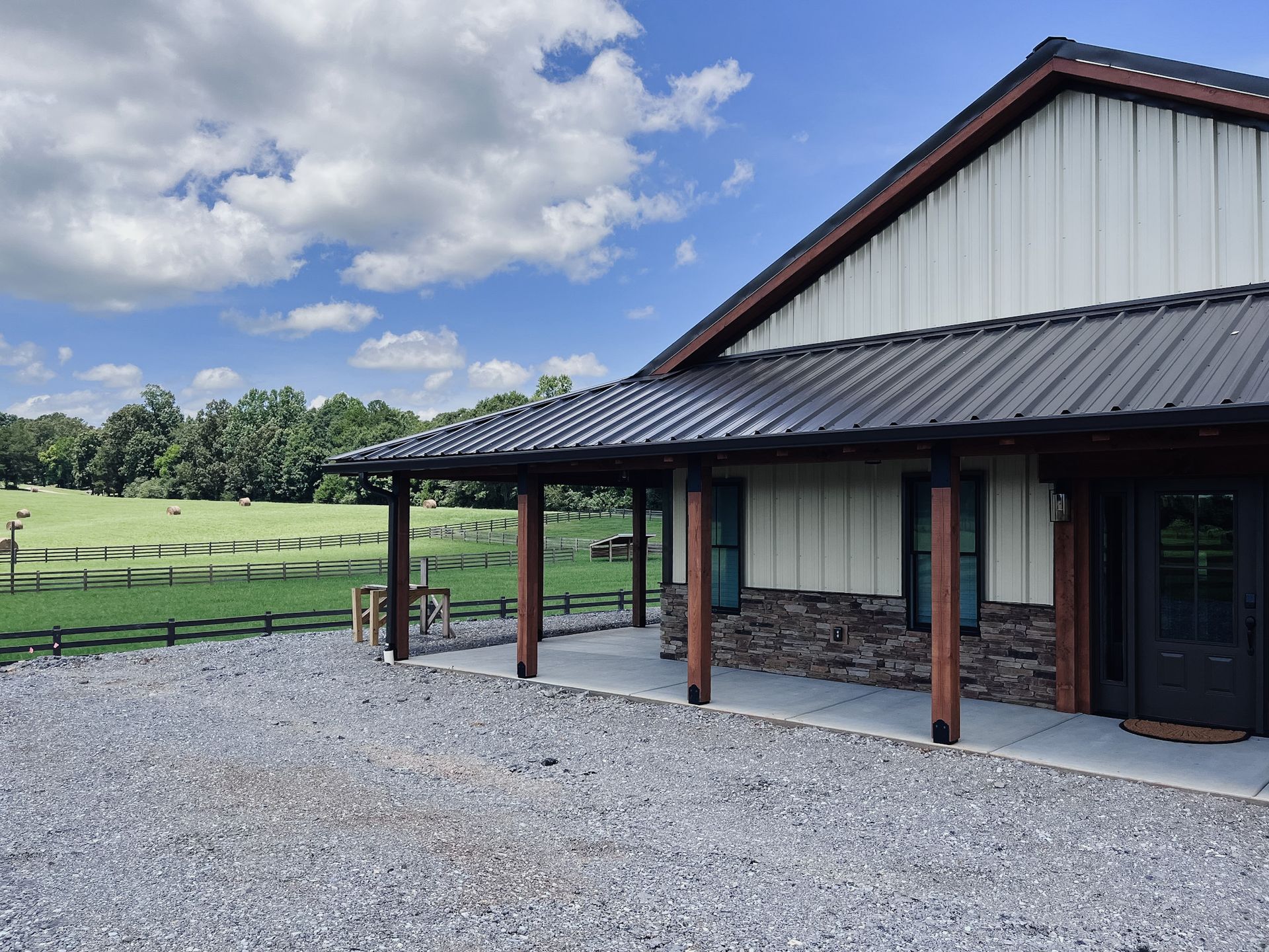 Rustic barn-style building with stone base, covered porch, gravel lot, and grassy field under a cloudy sky