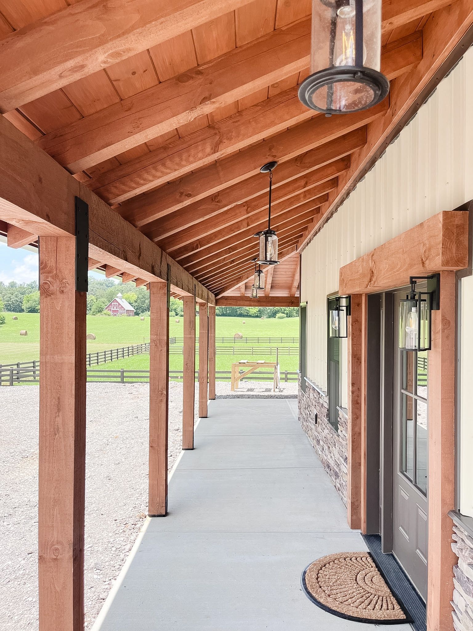 Covered wooden porch with timber posts, concrete floor, hanging lights, and glass doors overlooking a grassy field
