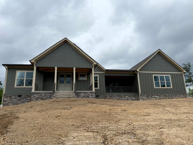 New gray house with stone base and front porch on a bare dirt lot under cloudy skies
