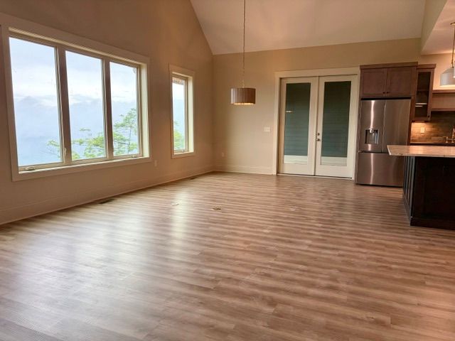 Empty open-plan living room with wood floors, large windows, and a kitchen area with stainless steel fridge.