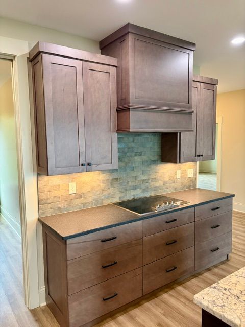 Modern kitchen with dark wood cabinets, beige countertop, mosaic backsplash, and stainless cooktop under a hood.