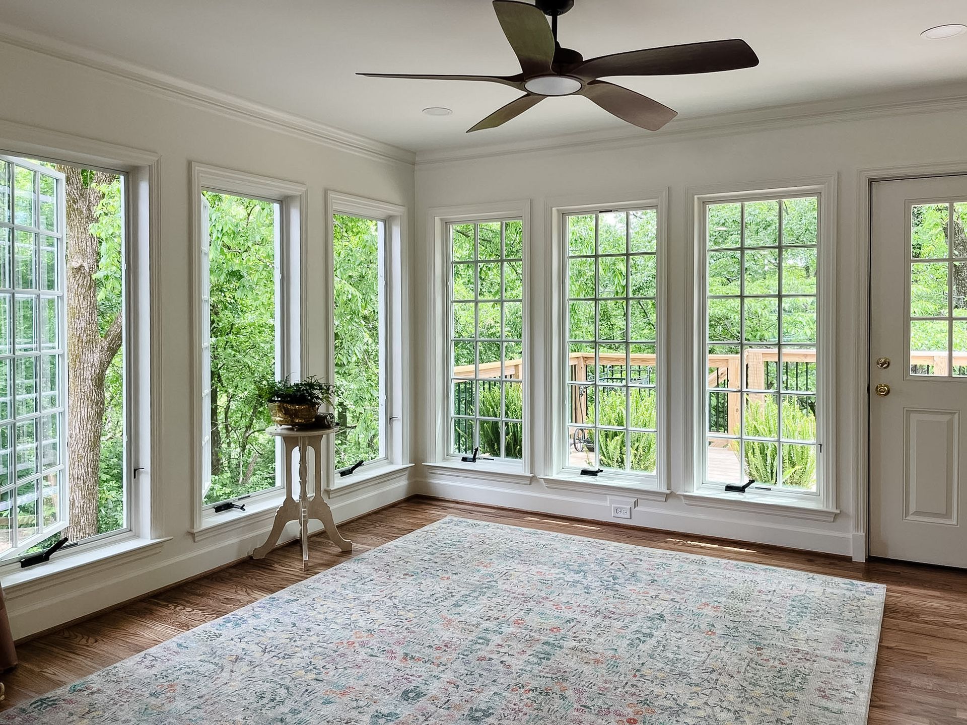 Bright sunroom with large windows, ceiling fan, patterned rug, and a potted plant on a stand