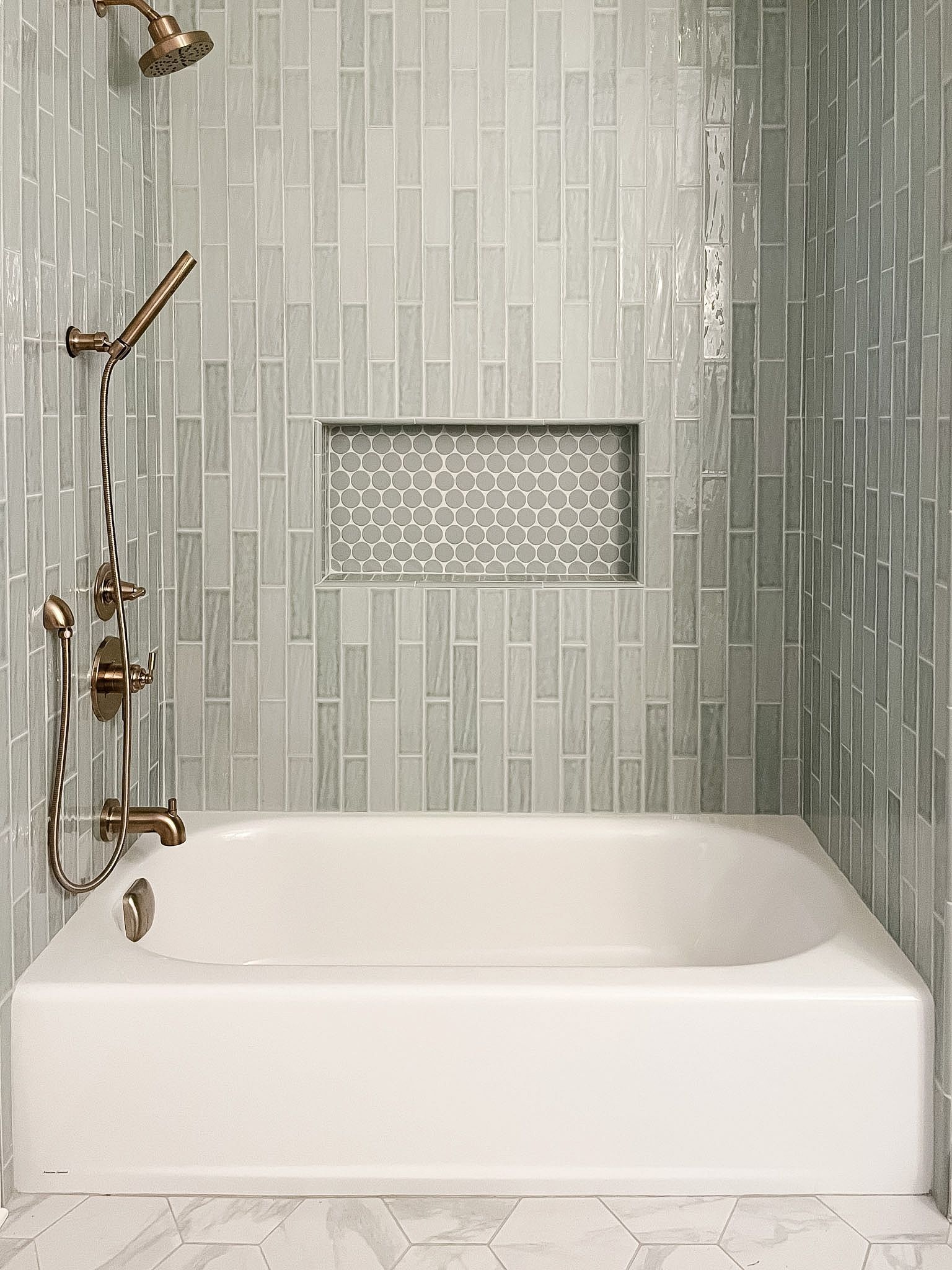 Modern white bathtub with bronze fixtures in a tiled gray bathroom, with a recessed shelf above it