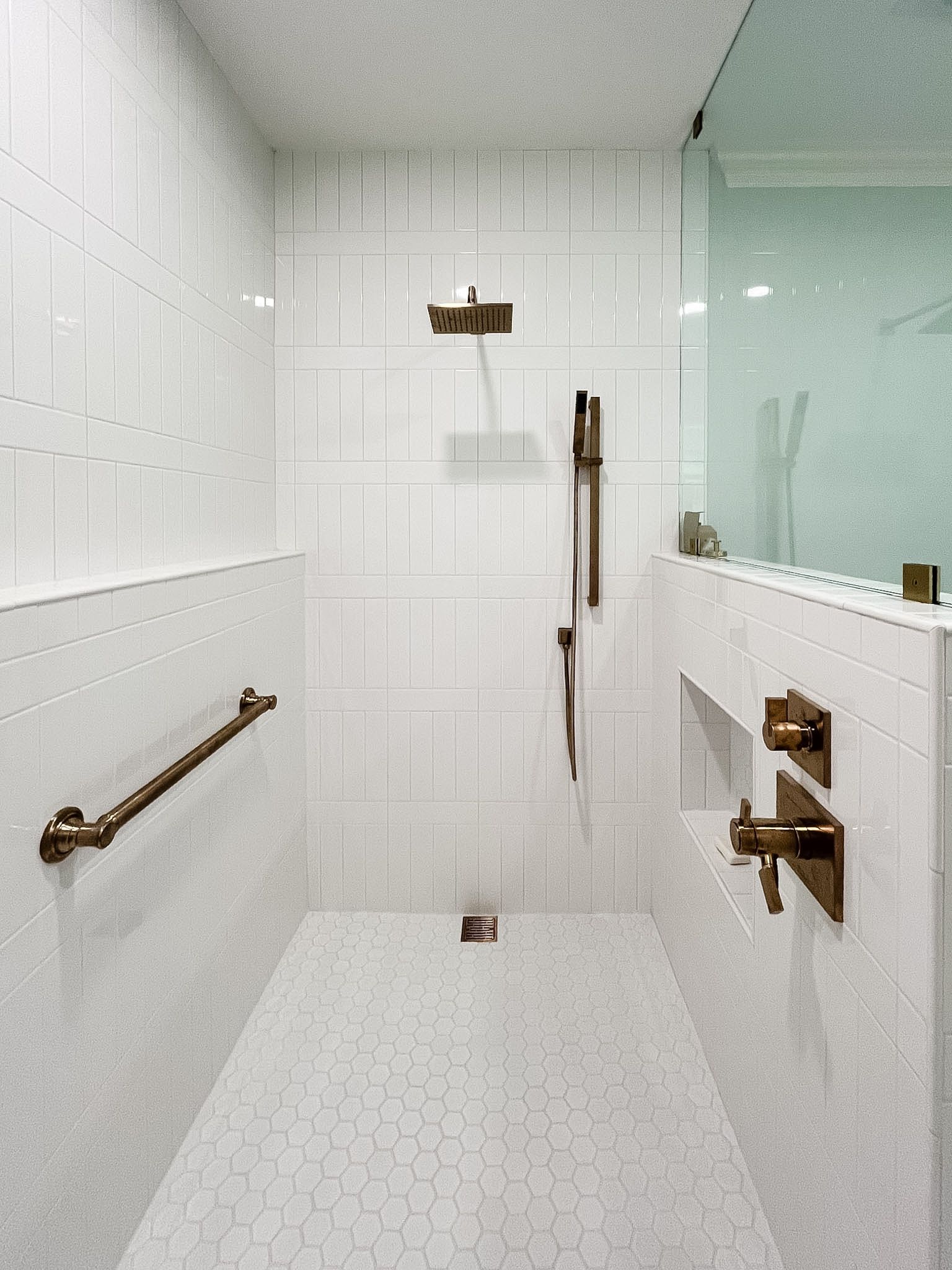 Modern white-tiled shower with brass fixtures, bench, grab bar, and glass partition