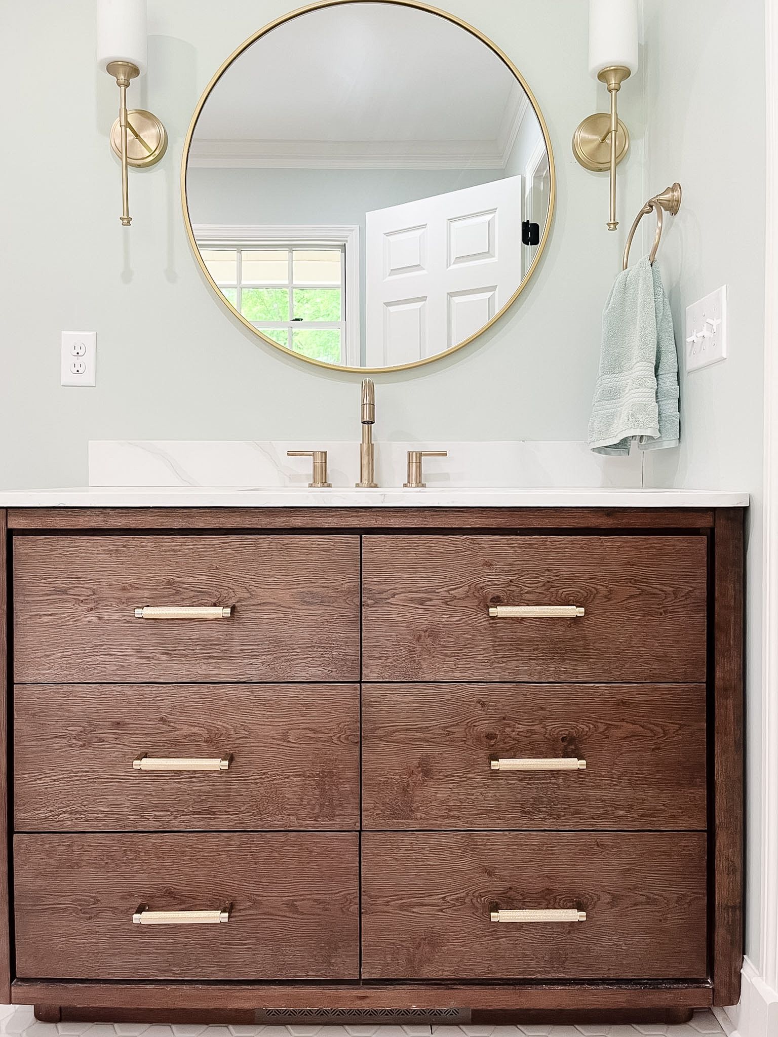 Modern bathroom vanity with brown wood drawers, round mirror, and brass sconces above a white sink