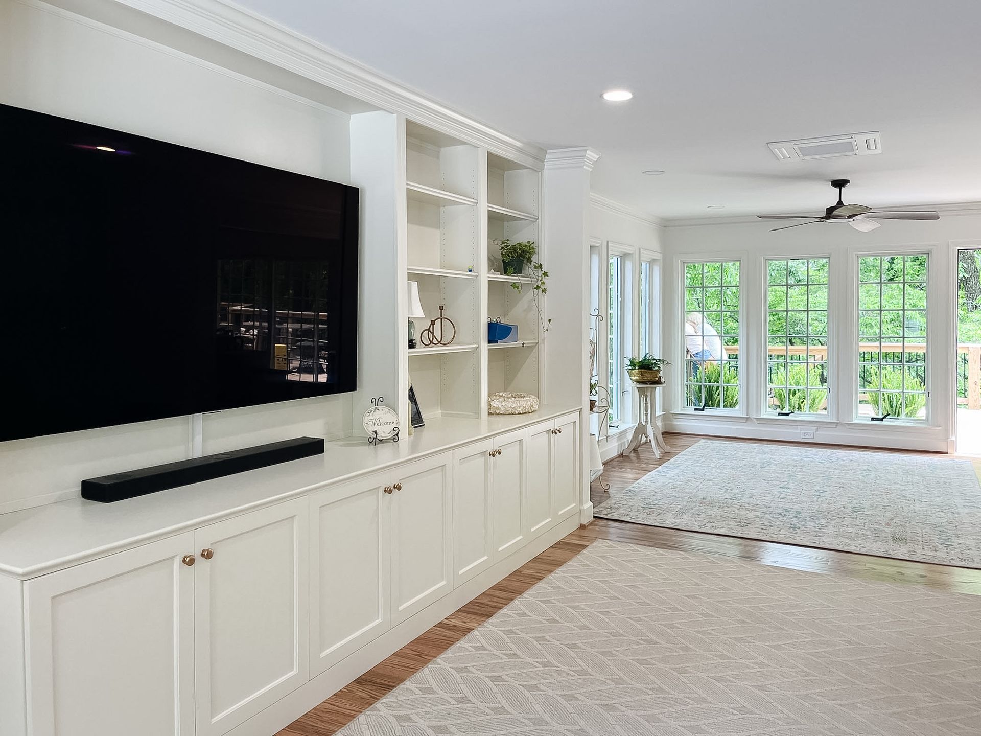 Bright living room with built-in white cabinets, large TV, and floor-to-ceiling windows overlooking greenery
