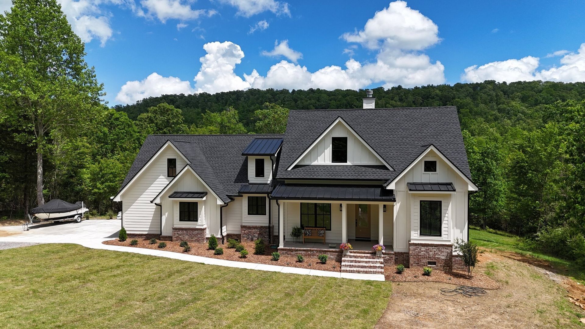 White house with black roof, brick accents, and green lawn against a forested hillside under a blue sky.