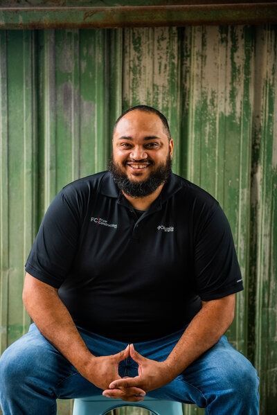 Smiling man in a black polo and jeans seated against a green corrugated metal wall