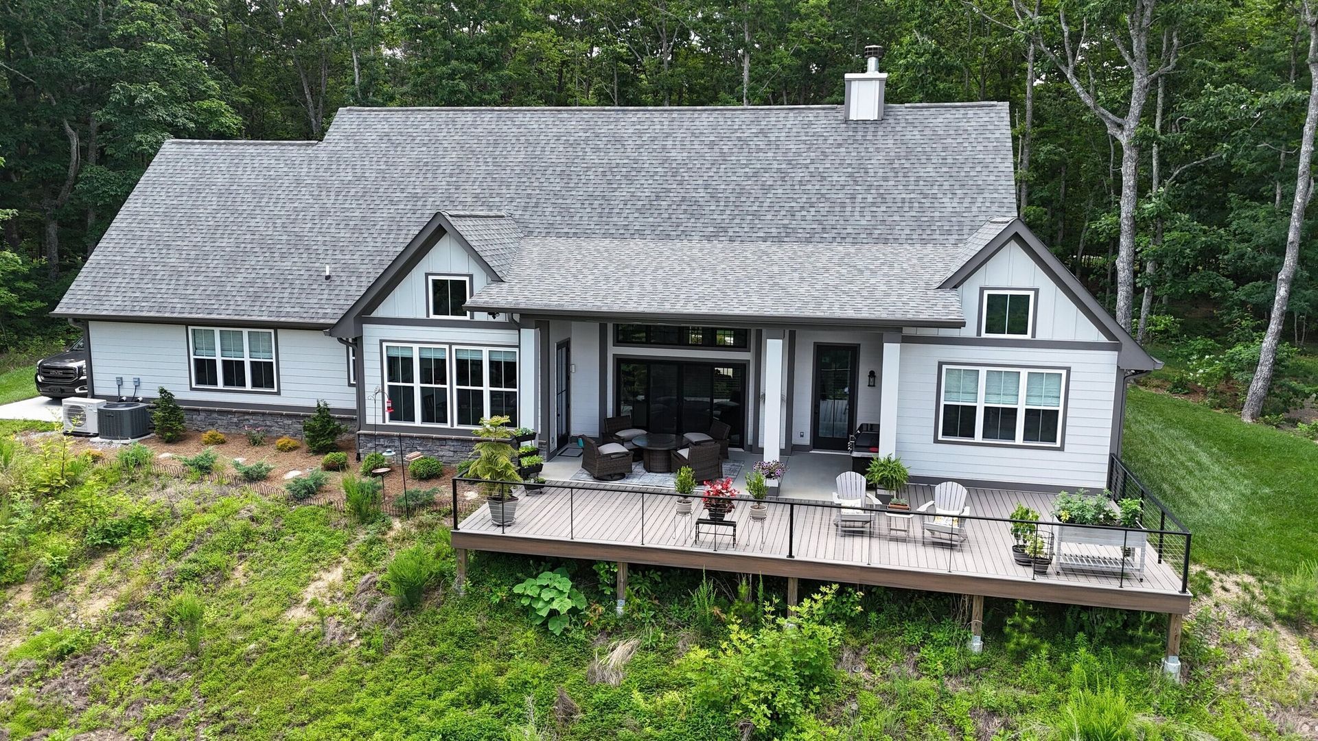 Gray house with a wooden deck, overlooking a grassy hill and forest.