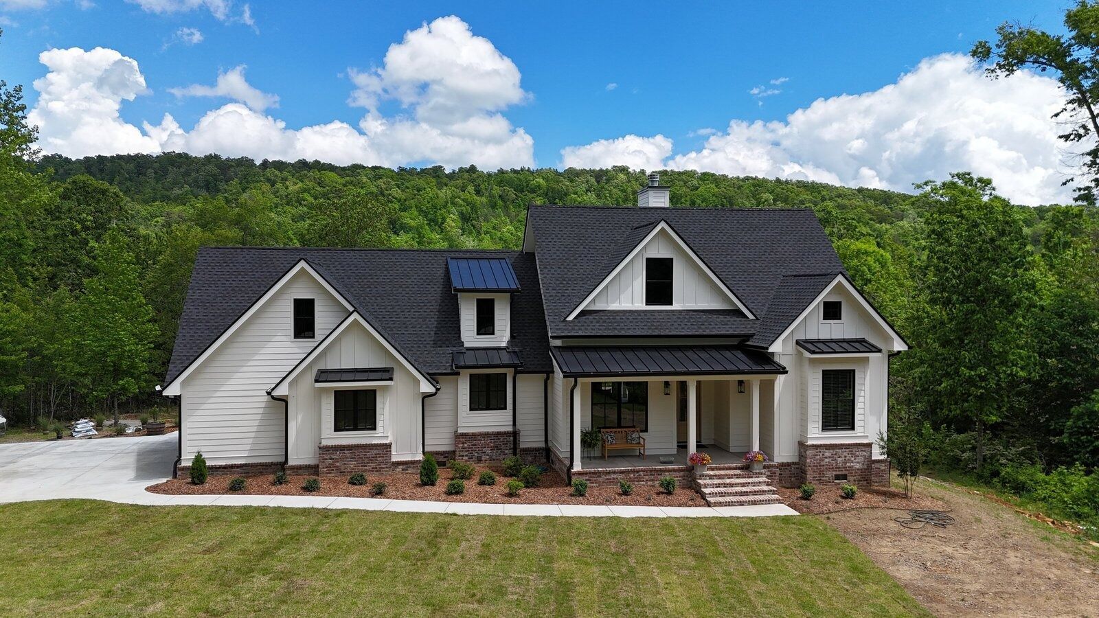 Farmhouse style home with gray roof, white siding, and stone accents, set against a backdrop of green trees and a blue sky.