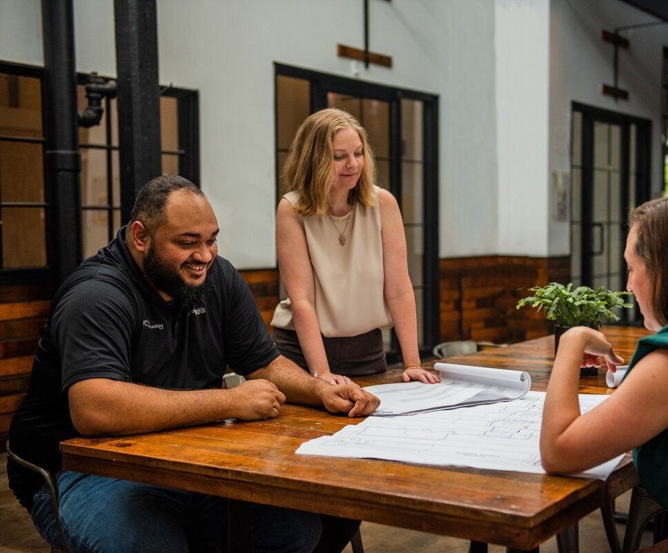 Three people lean over a large paper plan on a wooden table in a bright courtyard.