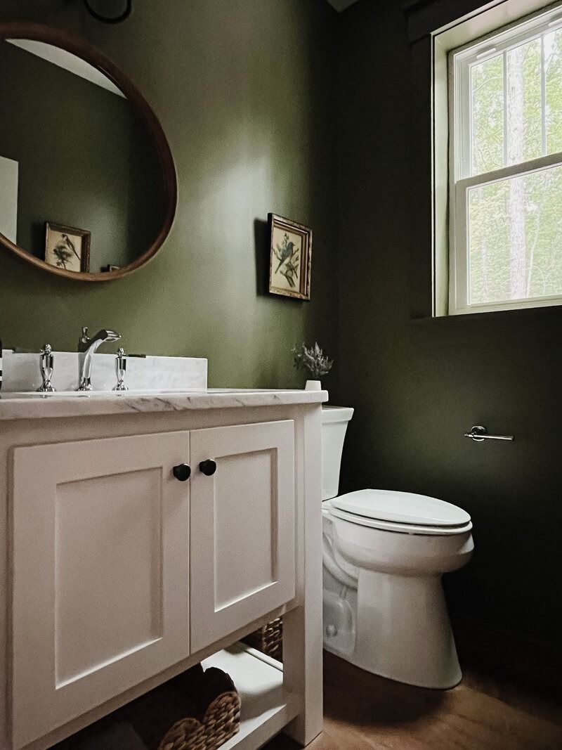 Small bathroom with dark green walls, white vanity and toilet, round mirror, and a window.